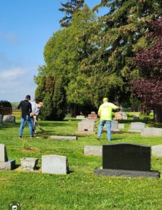 Volunteers cleaning cemetery grounds under sunny sky.